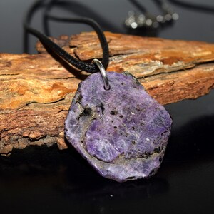 May include: A purple stone pendant on a black cord necklace. The pendant is an irregular shape with a rough texture and a silver-coloured bail. The stone has shades of purple and black.