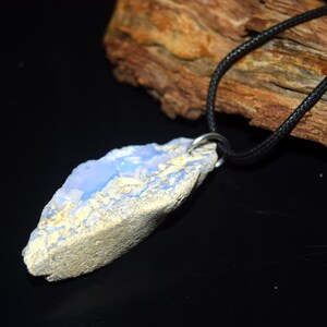 May include: A natural stone pendant with a light blue and beige color scheme. The pendant is suspended from a black cord necklace. The stone has a rough, textured surface and a unique, organic shape. The necklace is displayed against a dark background.
