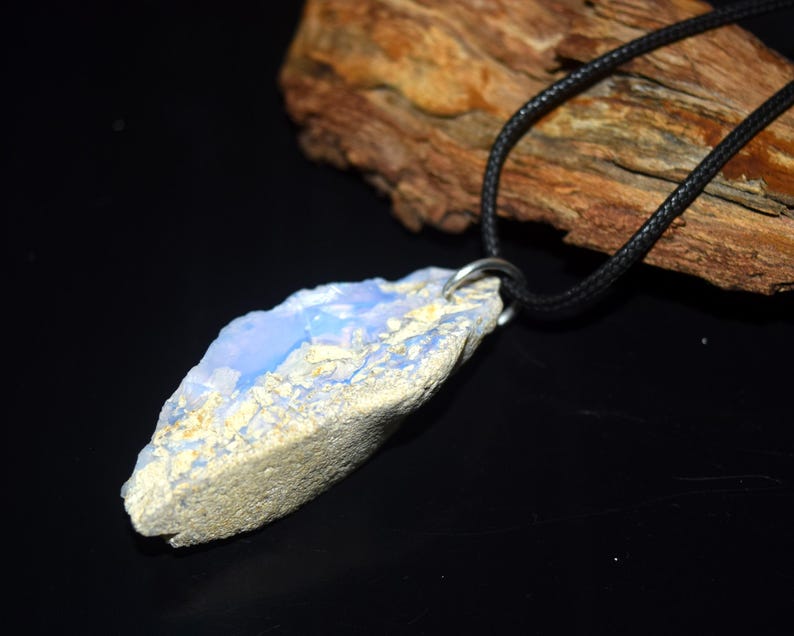 May include: A natural stone pendant with a light blue and beige color scheme. The pendant is suspended from a black cord necklace. The stone has a rough, textured surface and a unique, organic shape. The necklace is displayed against a dark background.