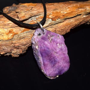 May include: A natural purple gemstone pendant on a black cord. The pendant has a rough, uneven shape and a textured surface. The cord is attached to the stone with a silver-coloured bail. The pendant is set against a backdrop of a piece of wood.