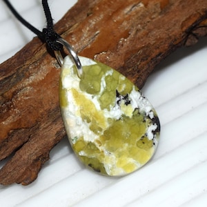 Peut inclure: Un pendentif en forme de goutte d'eau avec une pierre verte, jaune, blanche et noire. La pierre est attachée à une boucle en métal argenté et à un cordon noir. Le pendentif est posé sur un fond en bois.