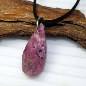 Puede incluir: Un collar con colgante de piedra preciosa de rubí en bruto en forma de lágrima. El rubí es de color rosa intenso con inclusiones negras y está suspendido de un cordón negro. El colgante está sujeto a un lazo de metal plateado.