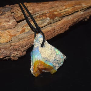 Puede incluir: Un colgante de ópalo en bruto sin pulir suspendido de un cordón negro. La piedra presenta una matriz gris rugosa y texturizada con destellos vibrantes de fuego iridiscente azul, verde y naranja en toda su superficie, mostrando formaciones minerales.