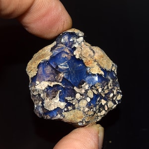 May include: A raw, irregular mineral specimen featuring a deep, translucent blue core with a glossy, glass-like texture. The stone is partially encased in a rough, beige, and grey rocky matrix with speckled, light-colored inclusions on one side.