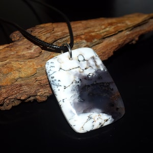 May include: A rectangular pendant featuring a polished white stone with dark, dendritic, tree-like inclusions and translucent grey patches. The stone is suspended from a simple black cord by a silver-toned metal bail, creating a necklace.