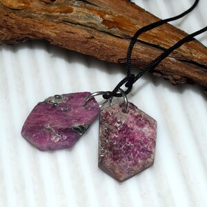 May include: Two raw ruby pendants in a dark pink hue, each with a unique, irregular shape. The pendants are suspended from a black cord necklace. The gemstones are attached to silver-coloured metal rings.