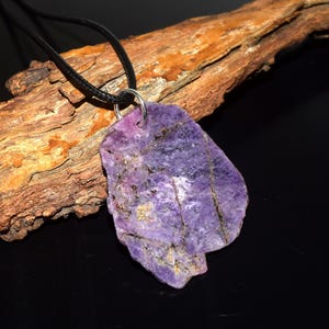 May include: A purple gemstone pendant on a black cord necklace. The stone has a mix of purple hues and natural patterns. The pendant is attached to a silver-coloured ring. The necklace is displayed on a wooden branch.