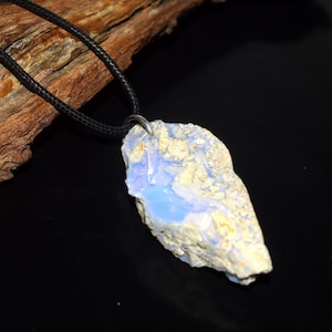 May include: A raw, light blue and cream-colored opal pendant hangs from a black cord necklace. The opal has a rough, natural texture and a polished, light blue center. The necklace is displayed against a dark background with a piece of wood.