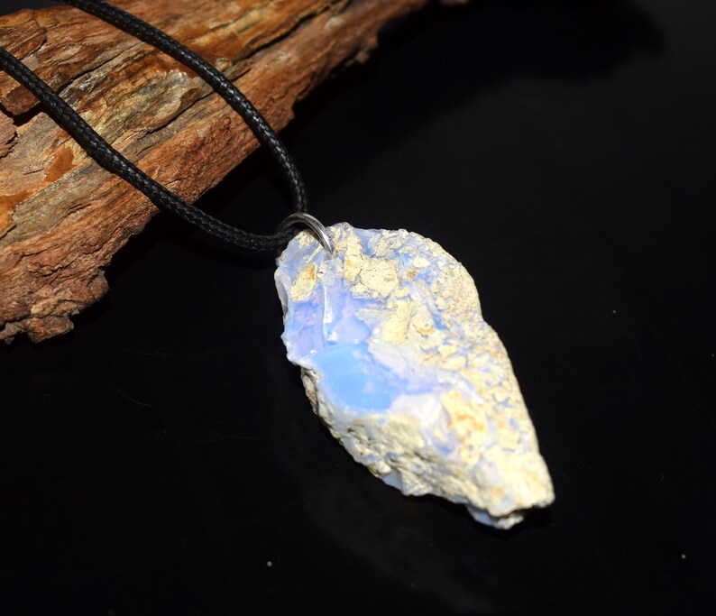 May include: A raw, light blue and cream-colored opal pendant hangs from a black cord necklace. The opal has a rough, natural texture and a polished, light blue center. The necklace is displayed against a dark background with a piece of wood.