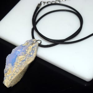 May include: A natural stone pendant necklace. The pendant is a rough-cut, light blue and beige stone. It hangs from a black cord necklace with a silver-tone clasp. The necklace is displayed on a white surface.