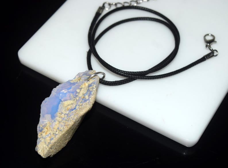 May include: A natural stone pendant necklace. The pendant is a rough-cut, light blue and beige stone. It hangs from a black cord necklace with a silver-tone clasp. The necklace is displayed on a white surface.