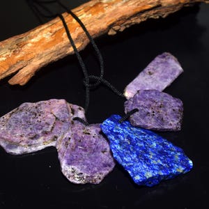 May include: A necklace featuring four irregular, flat, raw purple stone pendants and one vibrant blue, textured stone pendant, all suspended from a thin black cord. The stones have natural variations, dark speckles, and rough, unpolished edges.