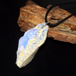May include: A pendant necklace featuring a large, irregularly shaped, light blue and cream-colored stone. The stone is suspended from a black cord. The pendant has a natural, raw appearance, with a textured surface and a silver-colored bail.