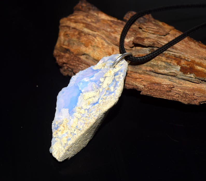 May include: A pendant necklace featuring a large, irregularly shaped, light blue and cream-colored stone. The stone is suspended from a black cord. The pendant has a natural, raw appearance, with a textured surface and a silver-colored bail.