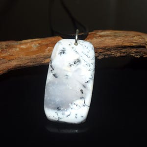 May include: A rectangular dendritic opal pendant featuring a smooth, polished white surface with intricate, fern-like black inclusions. The stone is suspended from a simple silver-toned metal bail attached to a thin black cord necklace.