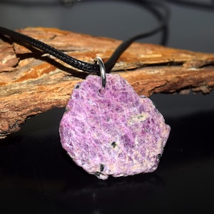May include: A raw, natural purple gemstone pendant on a black cord necklace. The pendant has an irregular shape, textured surface, and small black inclusions. The necklace features a silver-coloured bail.