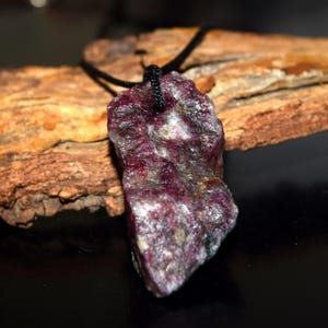 Puede incluir: Colgante de rubí natural en bruto con cordón negro. El rubí, de color rojo-púrpura intenso, tiene una superficie rugosa y texturizada. El colgante cuelga de un cordón negro, creando un collar sencillo y terroso.
