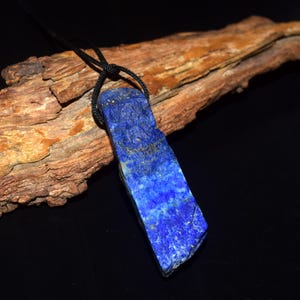 May include: A rectangular pendant made of raw, deep blue lapis lazuli stone with visible gold-colored pyrite flecks. The stone is suspended from a thin, black cord that loops through a hole at the top of the pendant, creating a simple necklace.