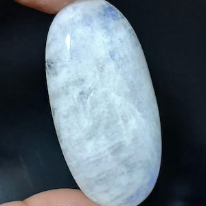 May include: Oval-shaped, polished gemstone with a milky white and pale blue color. The stone has a smooth surface and is held between fingers against a dark background. The gemstone is approximately 1.5 inches in length.