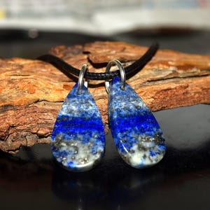 May include: Two teardrop-shaped pendants made of blue lapis lazuli, with silver-colored metal loops, hang from a black cord. The lapis lazuli has a striking mix of deep blue, gray, and white hues. The pendants are displayed against a natural wood background.