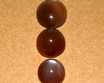 May include: Three polished, round gemstones in shades of brown and tan are arranged vertically against a neutral background. The stones have a smooth, reflective surface, with a subtle sheen. The gemstones appear to be cabochons, suitable for jewelry making.
