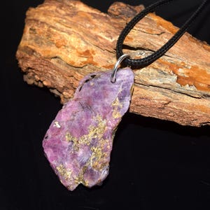 May include: A purple stone pendant necklace. The pendant is a rough-cut, irregular shape with gold-coloured inclusions. It hangs from a black cord with a silver-coloured metal loop. The necklace is displayed on a piece of wood.