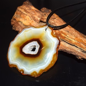May include: A pendant featuring a polished, circular slice of agate stone with concentric bands of white, amber, and dark brown, surrounding a central geode cavity with small white crystals, suspended from a thin black cord necklace.