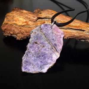 Könnte beinhalten: Eine Halskette mit einem Anhänger aus natürlichem lila Stein. Der Anhänger hat eine unregelmäßige, grob geschnittene Form mit Lavendel- und Violetttönen. Er hängt an einer schwarzen Kordelkette. Der Hintergrund umfasst ein Stück braunes Holz.
