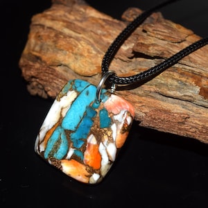 Pode incluir: Um colar com pingente retangular e cordão preto. O pingente apresenta um mosaico de pedra turquesa, laranja, branca e dourada. A pedra é presa a um anel de metal prateado.