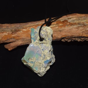 Puede incluir: Un colgante de ópalo en bruto sin pulir que presenta una matriz beige rugosa y texturizada con destellos iridiscentes azules y verdes. La piedra está suspendida de un cordón negro sencillo, mostrando su forma natural irregular y composición mineral.