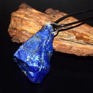 May include: A raw, deep blue lapis lazuli pendant with visible gold-colored pyrite flecks. The stone has an irregular, jagged shape and is attached to a thin black cord by a silver-toned metal jump ring, creating a simple necklace design.