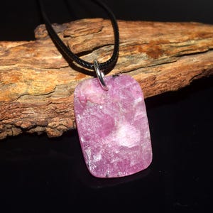 May include: A rectangular pink gemstone pendant with a textured, crystalline surface and a silver-toned metal bail, suspended from a thin black cord. The stone features natural variations in color and opacity, showcasing a vibrant magenta hue.