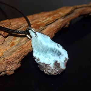 Puede incluir: Un colgante con una piedra Larimar en bruto en forma de lágrima de color azul claro con una base rocosa de color marrón oscuro texturizada. La piedra está unida a un sencillo enganche de metal plateado y cuelga de un fino collar de cordón negro.
