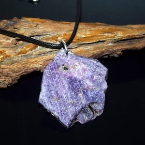Può includere: Un ciondolo di pietra preziosa viola grezza con una collana di cordino nero. La pietra di forma irregolare ha una superficie strutturata ed è sospesa da un'ansa color argento. Il ciondolo è esposto su uno sfondo scuro con un pezzo di legno.