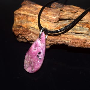 Peut inclure: Un pendentif en pierre précieuse rose en forme de goutte avec des inclusions sombres, suspendu à un cordon noir. La pierre présente une surface polie avec une texture naturelle et est fixée au cordon par une bélière en métal argenté.