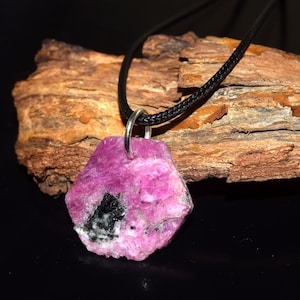 Puede incluir: Un colgante de rubí rosa en bruto con una inclusión negra, suspendido de un collar de cordón negro. La gema tiene forma irregular y tiene una fianza de color plateado. El fondo presenta un trozo de madera.