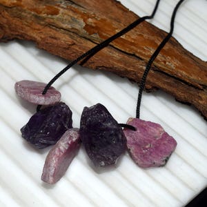 Pode incluir: Um colar com pedras preciosas brutas e não cortadas em tons de roxo e rosa. As pedras são enfiadas em cordões pretos e são exibidas sobre um fundo branco com um pedaço de madeira.