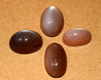 May include: Four polished, oval-shaped gemstones in varying shades of brown and tan are arranged on a light brown surface. The stones have a smooth, glossy finish, reflecting light. The gemstones are likely for jewelry making.