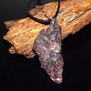 May include: A pendant necklace featuring a raw, natural stone. The stone is a mix of dark grey, purple, and white, with an irregular shape. It's suspended from a black cord. The pendant is set against a backdrop of textured wood.