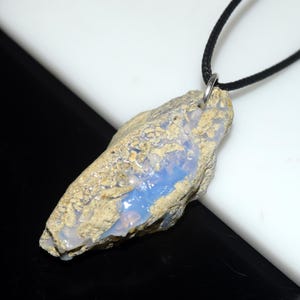 May include: A natural opal pendant with a black cord. The opal has a rough, textured exterior with a mix of tan and gray tones. A vibrant blue color is visible within the stone. The pendant is attached to a black cord with a silver-colored metal loop.