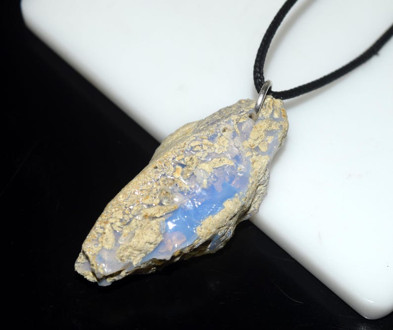 May include: A natural opal pendant with a black cord. The opal has a rough, textured exterior with a mix of tan and gray tones. A vibrant blue color is visible within the stone. The pendant is attached to a black cord with a silver-colored metal loop.