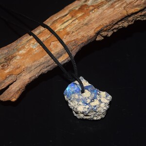May include: A raw, unpolished opal pendant featuring a rough, textured grey stone matrix with vibrant blue iridescent flashes. The stone is suspended from a simple black cord, showcasing the natural, irregular shape of the mineral specimen.