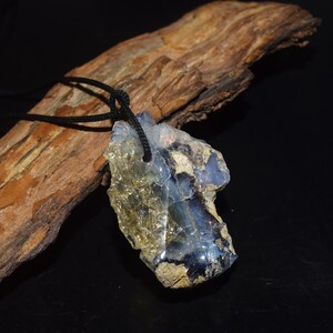 May include: A raw, irregular pendant featuring a translucent yellow and blue mineral stone with rough, unpolished edges. The stone is suspended from a thin black cord, showcasing a natural, jagged texture with visible crystalline layers.