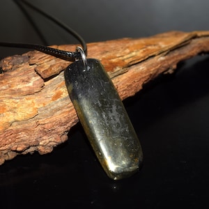 Könnte beinhalten: Ein rechteckiger Anhänger mit einer polierten, dunkelmetallischen Oberfläche und subtilen goldbraunen Untertönen. Der Anhänger hängt an einem dünnen schwarzen Band mit einem silberfarbenen Metallring und hat eine glatte, reflektierende Oberfläche.