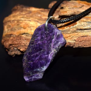 May include: A purple amethyst pendant with a silver bail, on a black leather cord. The gemstone has a rough, textured surface and varying shades of violet. The background features a piece of weathered wood.