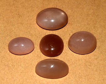 May include: Five polished, oval and round gemstones in shades of brown and pink. The gemstones are smooth and glossy, with a slight translucence. The arrangement includes a dark brown, circular stone and four lighter, oval-shaped stones.
