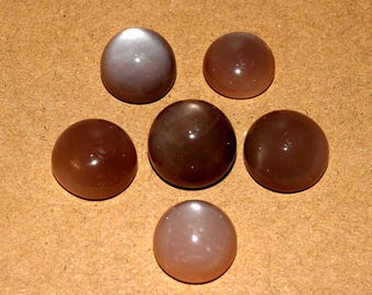 May include: Six cabochon gemstones in shades of brown and gray are arranged on a light brown surface. The stones have a smooth, rounded shape and a polished, reflective surface. The colors range from light tan to dark brown.