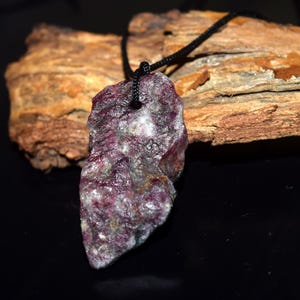 Peut inclure: Pendentif en rubis naturel brut et non taillé. La pierre précieuse est d'un violet profond avec des inclusions blanches et grises, suspendue à un cordon noir. Le fond présente du bois texturé et une surface sombre.