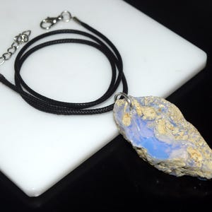 May include: A natural opal pendant necklace. The pendant is a rough-cut stone with a blue and beige color scheme. The necklace has a black cord with a silver-colored clasp and chain.