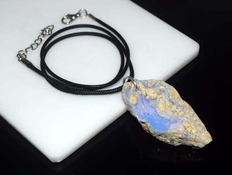 May include: A natural opal pendant necklace. The pendant is a rough-cut stone with a blue and beige color scheme. The necklace has a black cord with a silver-colored clasp and chain.
